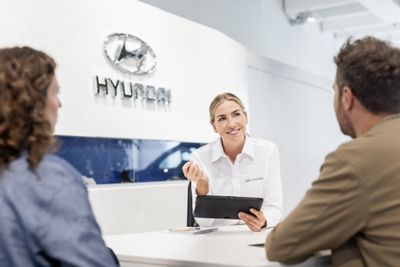 Eine Mitarbeiterin eines Hyundai Autohauses mit einem Tablet in der Hand berät eine Frau und einen Mann an ihrem Schreibtisch.