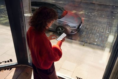 Woman standing by the window, looking at the Bluelink app on her phone. Her Hyundai i30 Fastback is parked on the street. 