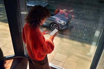 Eine Frau mit Smartphone blickt durch ein Fenster auf ihren vor dem Haus geparkten Hyundai i20.