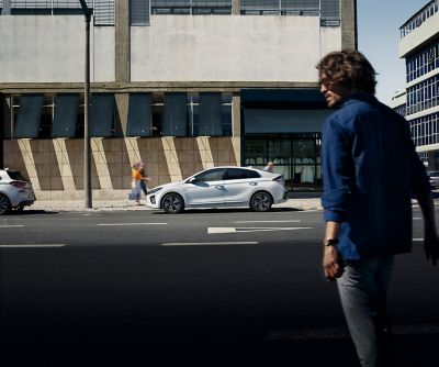 A man about to cross the road to get to his parked Hyundai IONIQ. He holds a smartphone in his hand.