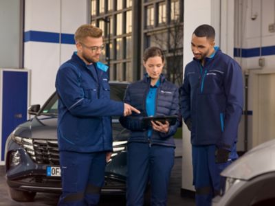 Traja technici Hyundai v modrých uniformách pozerajúci na tabuľku v dielni.