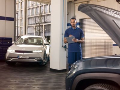 Hyundai mechanic stands in the workshop with a clipboard and looks at the car.