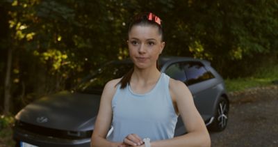 Bezkyna v modrom tielku a s bezeckymi okluarmi na hlave stojaca pred Hyundai IONIQ 5 sivej farby, pripravujuca sa na beh. Pravu ruku ma pripravenu na tlacidle smart hodiniek pripravujuc sa na spustenie behu