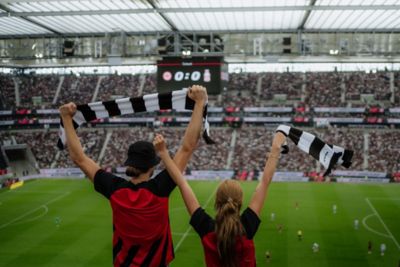 Zwei Eintracht Frankfurt Fans jubeln mit Fan-Schals von der Tribüne in einem Fußballstadion.