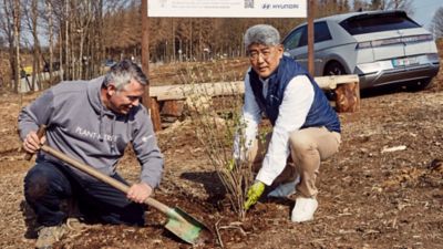 Der Hyundai Präsident Wang Chul Shin pflanzt zusammen mit einem Mitarbeiter einen Baum im IONIQ Wald. Im Hintergrund steht eine Hyundai IONIQ 5.
