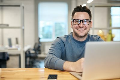 Ein lachender Mann mit Brille sitzt an einem Schreibtisch mit Laptop und Smartphone.