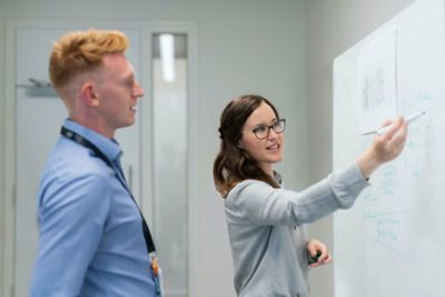 Eine Frau zeigt mit einem Stift auf ein Whiteboard, ein Mann steht dahinter und sieht zu.