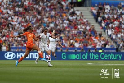 Twee vrouwelijke voetballers op het veld met op de achtergrond het reclamebord Hyundai Goal of the Century.