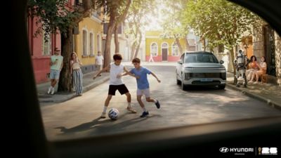 Zwei Kinder spielen Fußball auf einer ruhigen Straße, während ein modernes Hyundai-Auto und ein humanoider Roboter am Straßenrand stehen; Szene aus einer Kampagne zur Kooperation mit der FIFA 2026.