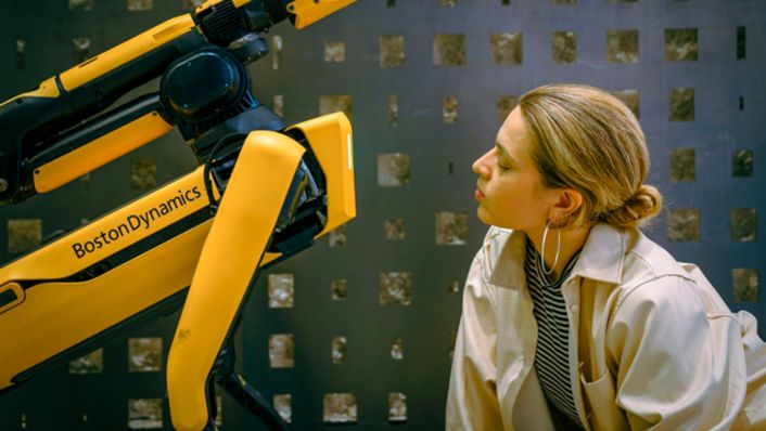 A woman is looking at the Boston Dynamics robot Spot, symbolizing how advances in AI research help make robots easier to use.