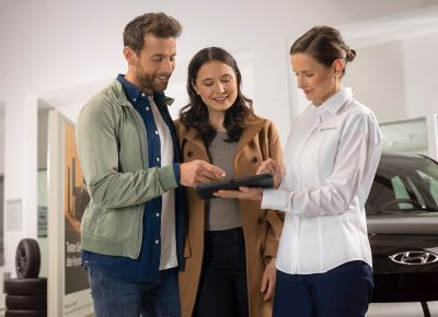 Three people stand in a car dealership, two looking at a tablet held by a third person in a white shirt with a logo. A black car is partially visible in the background.