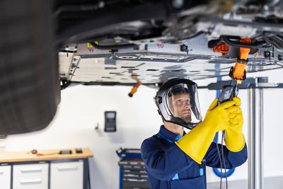 A mechanic wearing a protective face shield and yellow gloves is working underneath a vehicle on a lift, using an orange power tool.