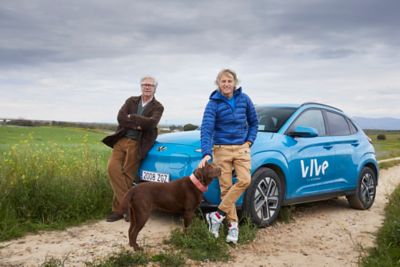 Fotografía con vecino de Illán de Vacas y Jesús Calleja acariciando un perro apoyados sobre un KONA Eléctrico aparcado en un camino rural rodeado de vegetación.