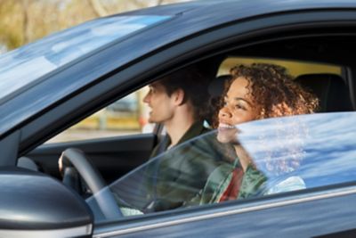 A woman and man sit in a Hyundai i20 with the driver's side window half down, outside view