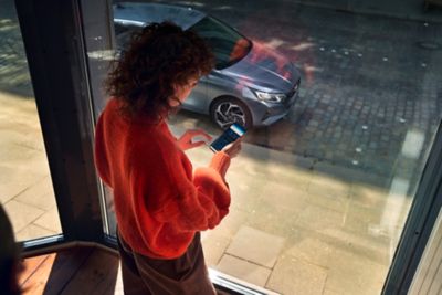 Una mujer mirando la aplicación de Hyundai Bluelink desde su smartphone, con su Hyundai i20 aparcado en la calle.