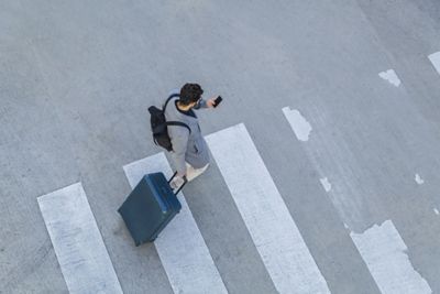 A man in a crosswalk pulling a suitcase and looking at his phone.