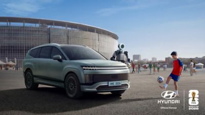 Hyundai SANTA FE parkerad utanför en fotbollsstadion tillsammans med Boston Dynamics-roboten Atlas och ett barn som spelar fotboll under FIFA World Cup 2026™-kampanjen.