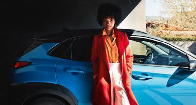 A woman in a long red coat, leaning against the passenger door of her Hyundai KONA Hybrid.