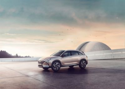 The Hyundai Nexo, shown from the side, standing before a futuristic building at sunset.
