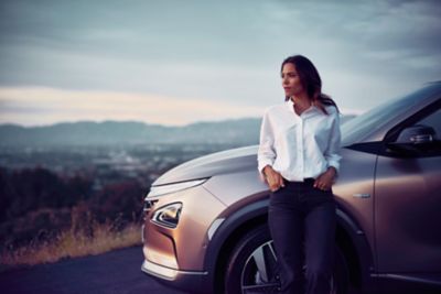 Eine Frau in weißer Bluse lehnt seitlich an der Motorhaube des Hyundai NEXO, im Hintergrund ein Landschaftspanorama.