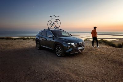 Ein Mann steht neben seinem Hyundai TUCSON mit Fahrrad-Dachträger und schaut aufs Meer.