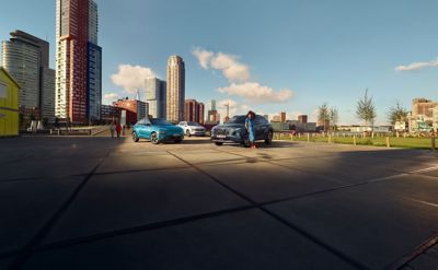 Drei Hyundai Modelle parken auf einem Platz vor einer Großstadt. An einem Fahrzeug lehnt eine Frau in blauem Kleid.
