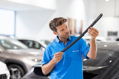 Three people stand in a car dealership, two looking at a tablet held by a third person in a white shirt with a logo. A black car is partially visible in the background.