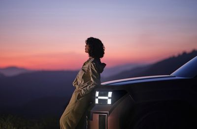 Hyundai IONIQ 9’un arka koltuğunda fotoğraf makinesiyle oturan bir kişi, açık kapıdan mor-pembe renkteki gökyüzü görünüyor. 