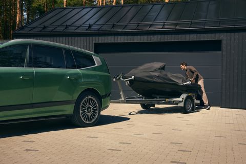 A Hyundai IONIQ 9 is parked in front of a garage. A covered boat trailer is attached to the vehicle’s tow hitch.