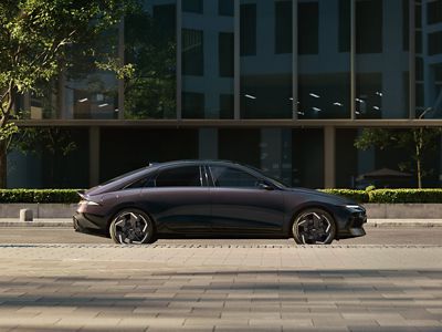 Elektrický sedan Hyundai IONIQ 6 zobrazený z úrovně ulice, zaparkovaný s moderní budovou v pozadí.