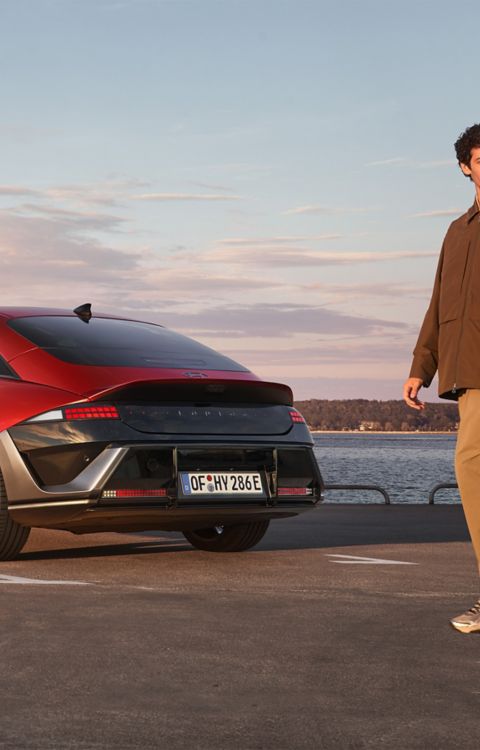 A man and a woman walking past a red Hyundai IONIQ 6 N Line parked next to the ocean.