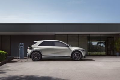 Boční pohled na nový IONIQ 5 v šedé barvě, připojený k nabíjení mimo šedou budovu.