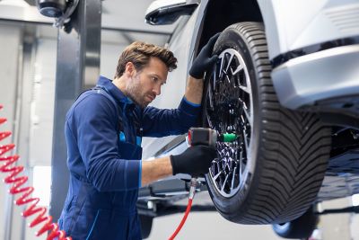 A mechanic in a blue uniform and black gloves uses a power tool on a car wheel lifted on a hydraulic lift.