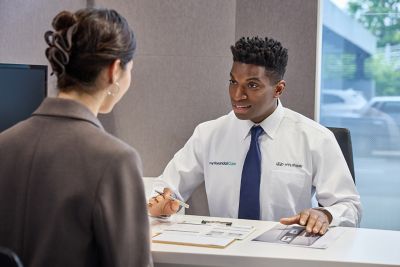 A woman is sitting across from a man in a white shirt and blue tie at a desk, discussing documents.