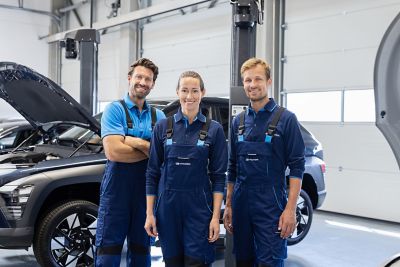 Three mechanics in blue uniforms stand in a garage with two cars on lifts behind them.