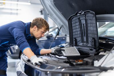 Mechanic in blue uniform inspecting a car engine while holding an air filter.