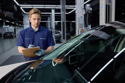 Mechanic in blue uniform inspecting a car windshield with a clipboard in hand.